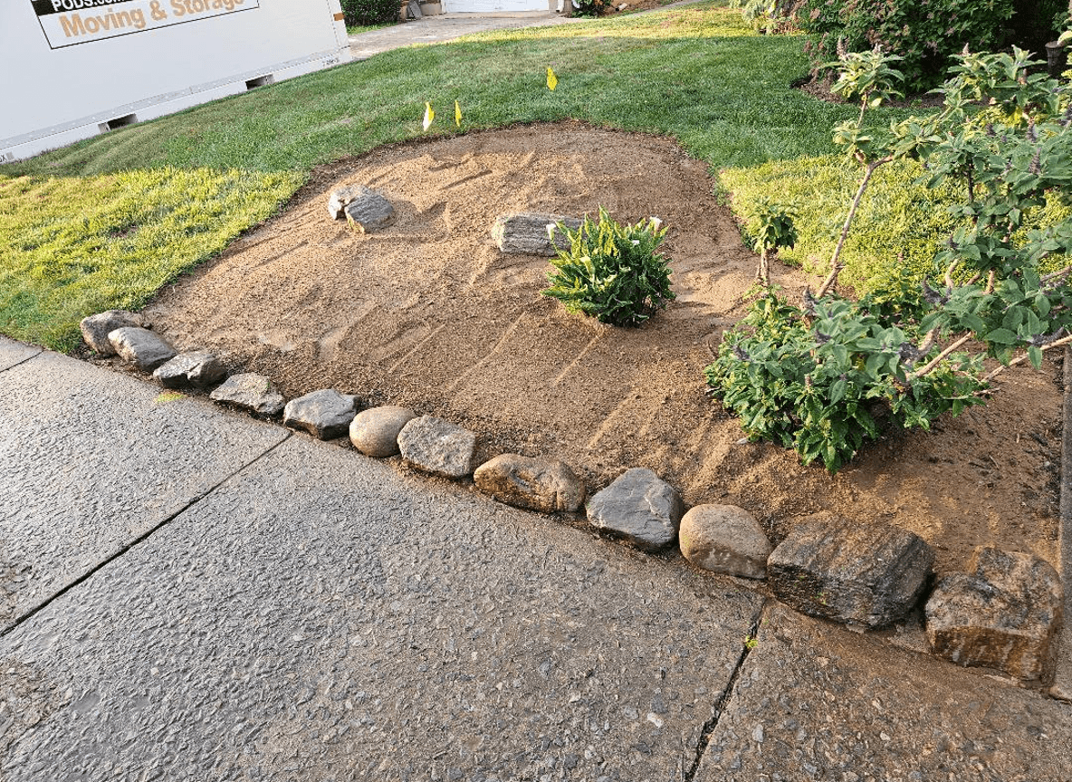 A garden area with rocks bordering a sidewalk, preparing for perennial planting.