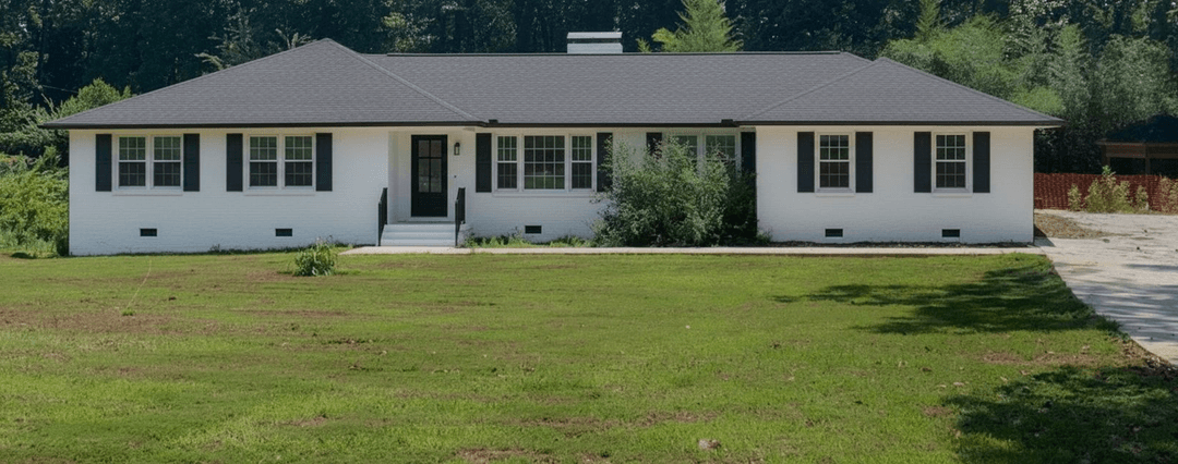 A front yard with a house, featuring a large grassy area and some overgrown shrubs.