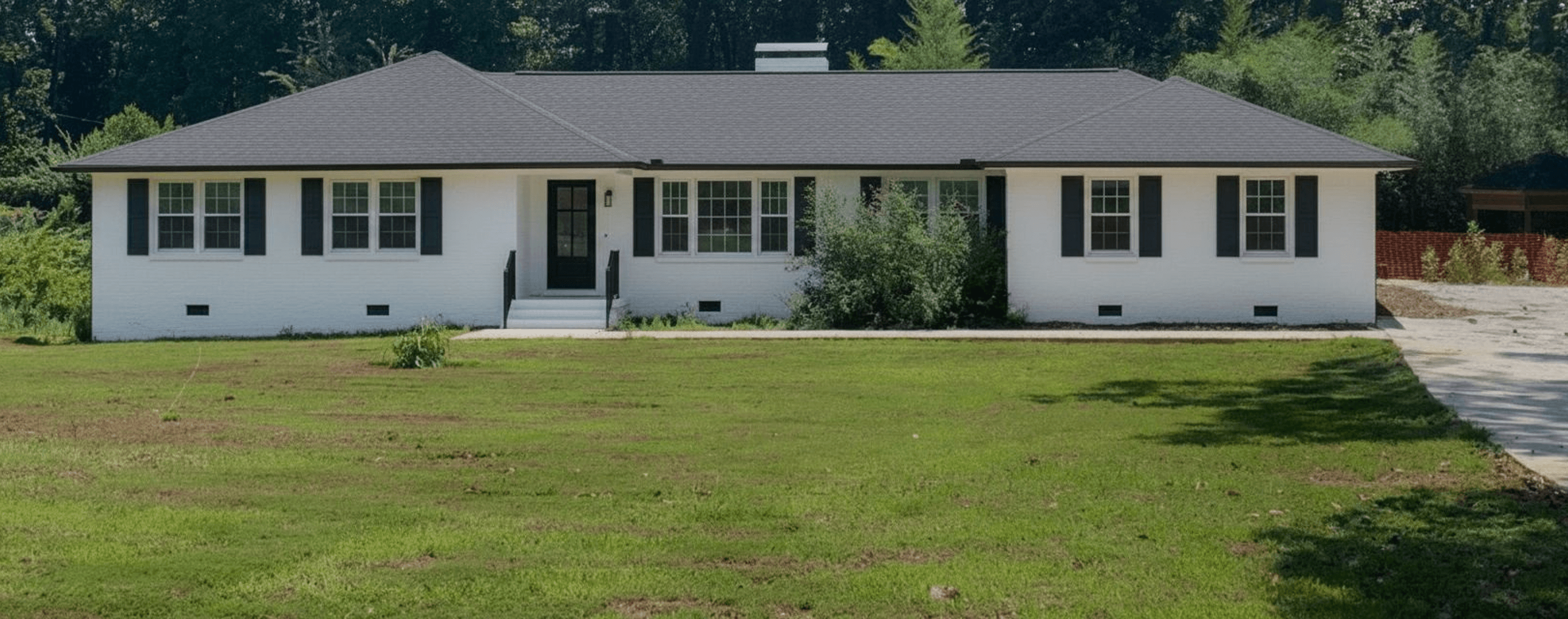 A front yard with a house, featuring a large grassy area and some overgrown shrubs.