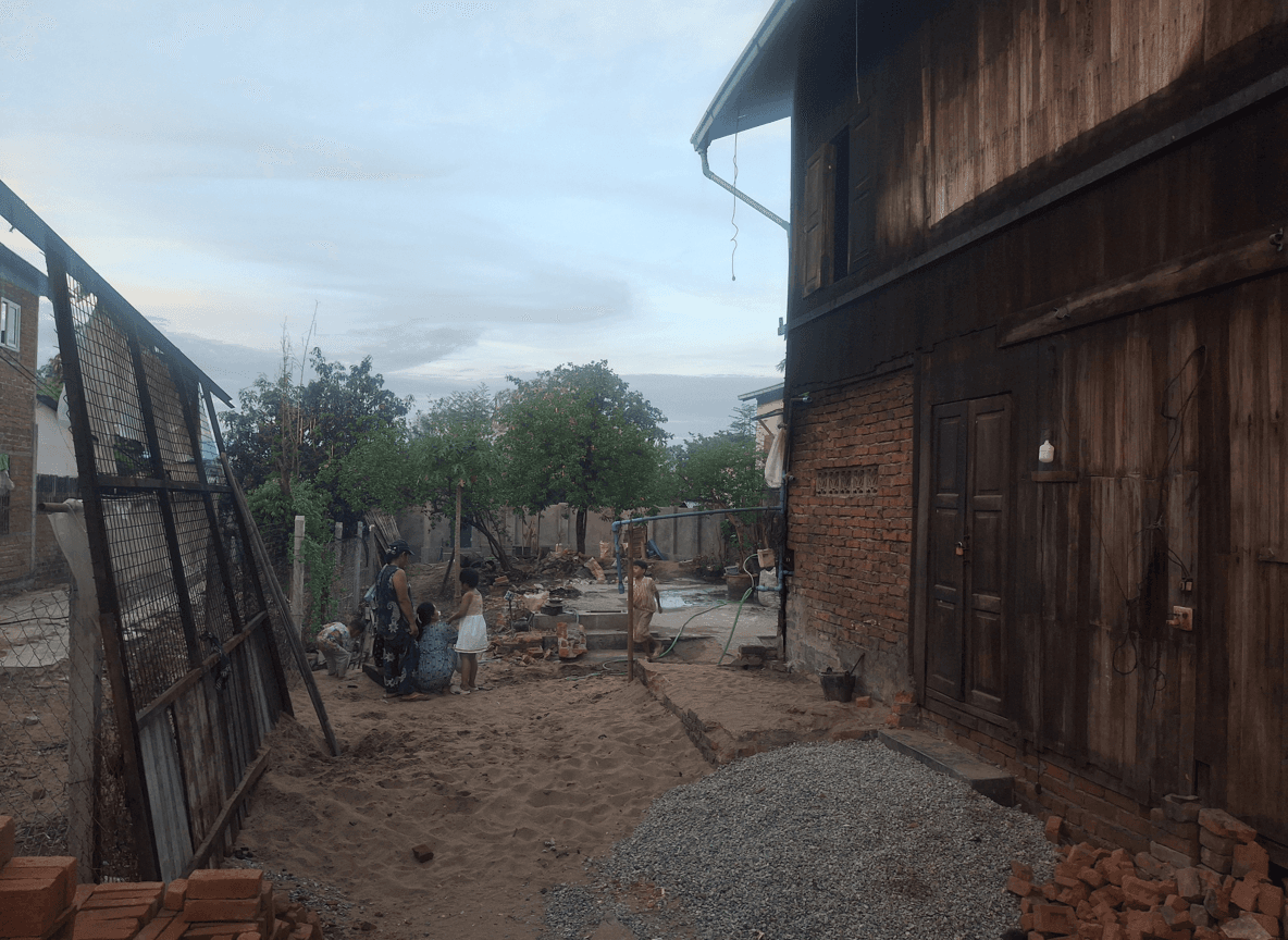 A sandy yard with people working on landscaping, surrounded by trees and a wooden house.