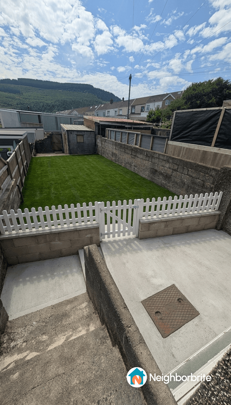 A view of a grassy yard with a white picket fence and surrounding structures.