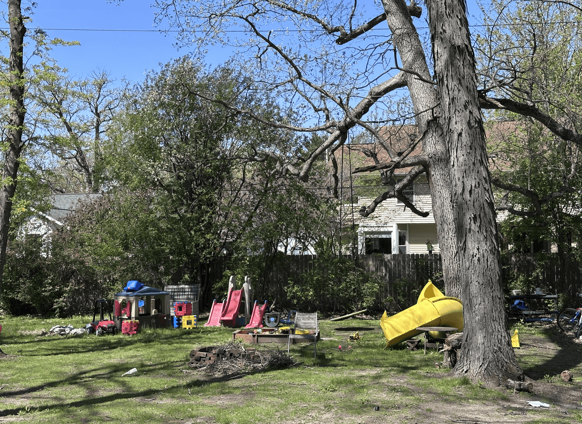 A suburban backyard with a swingset, slide, and playhouse under trees.