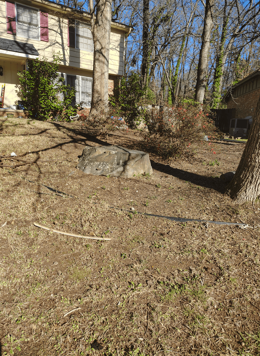 A large rock on a slight hill in a yard with trees and shrubs.